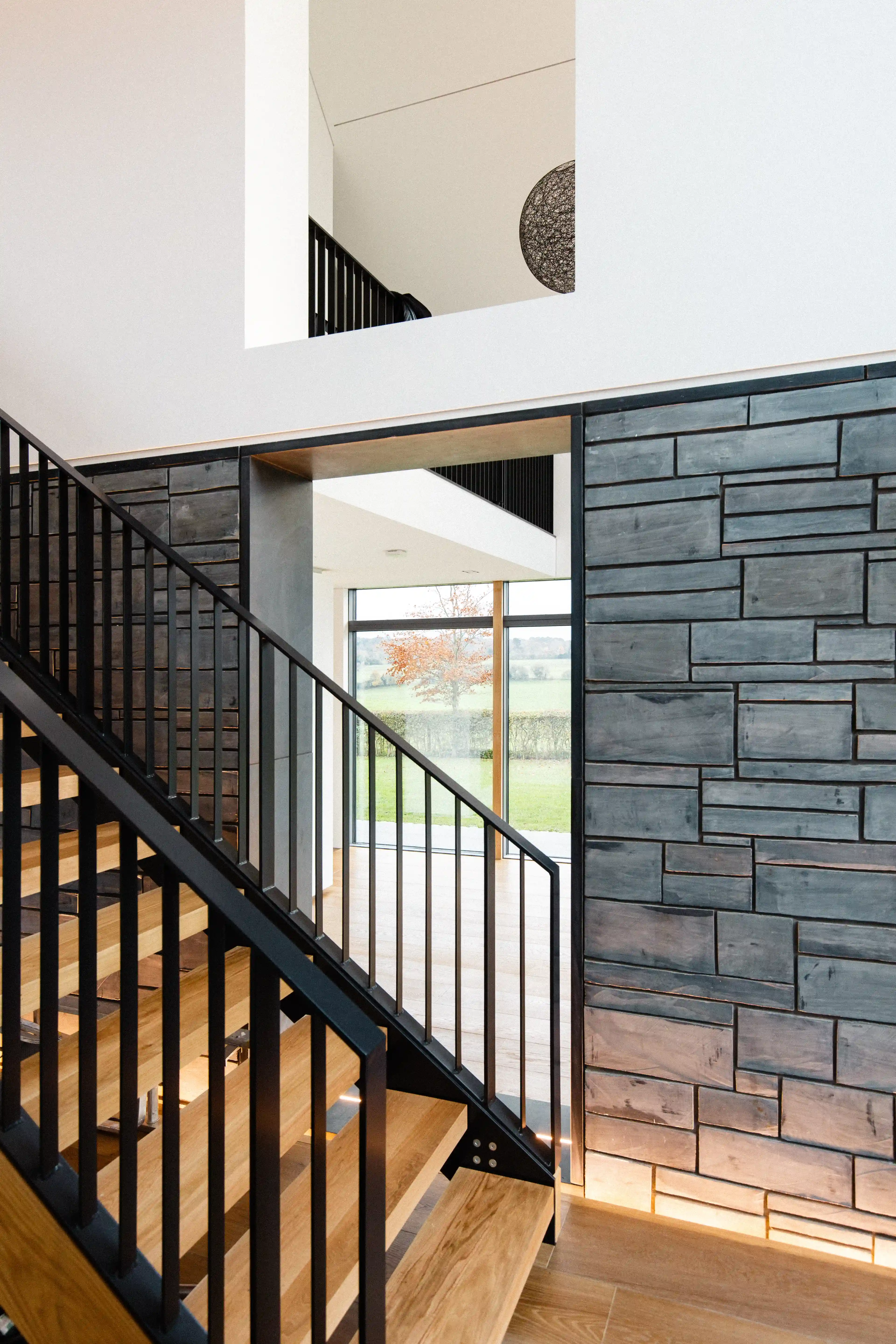 Double-height stair hall with oak treads, black steel balustrade and a dark stone wall, looking through to the fields