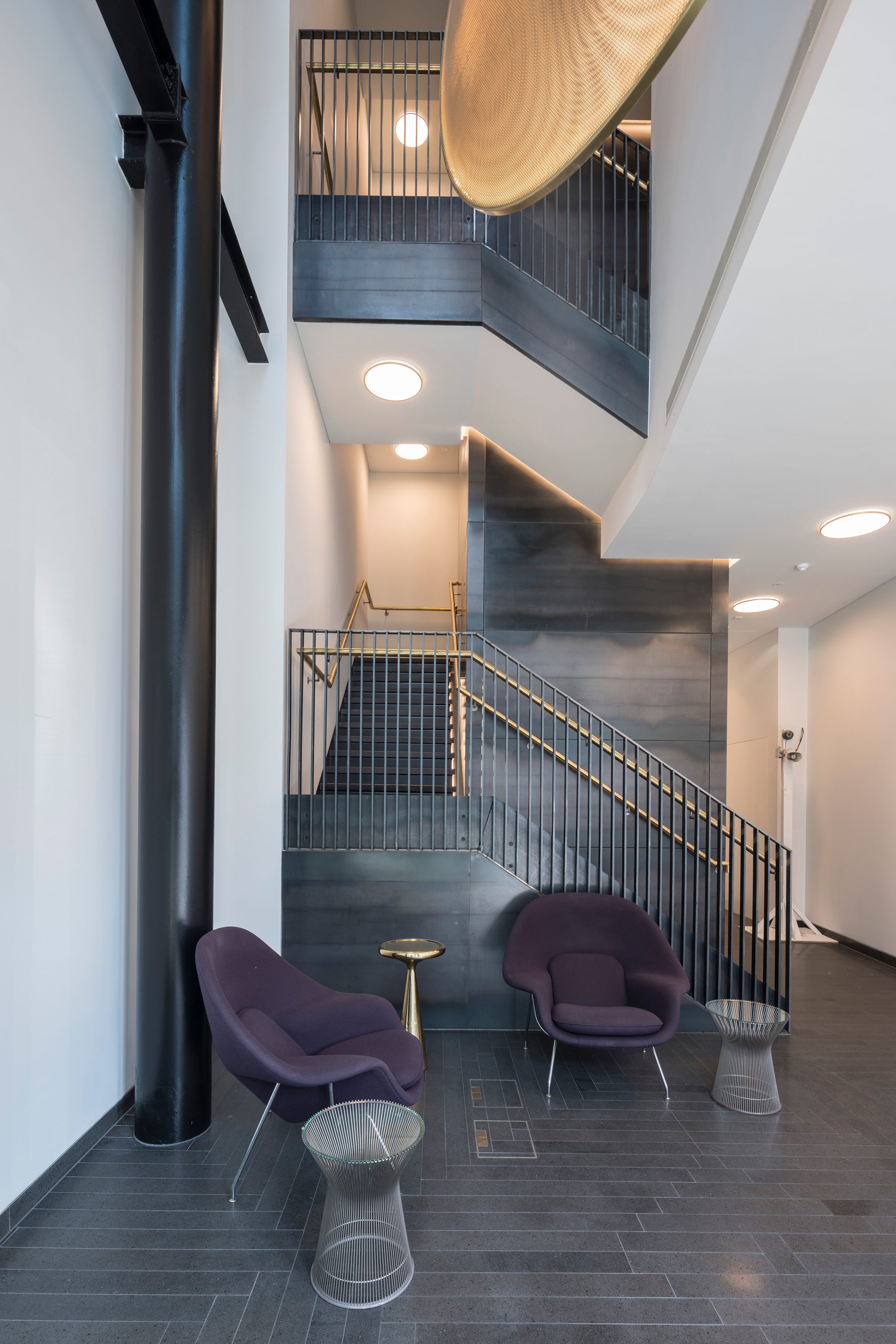 Reception with the stair rising past dark metal cladding, brass handrails and a large perforated pendant