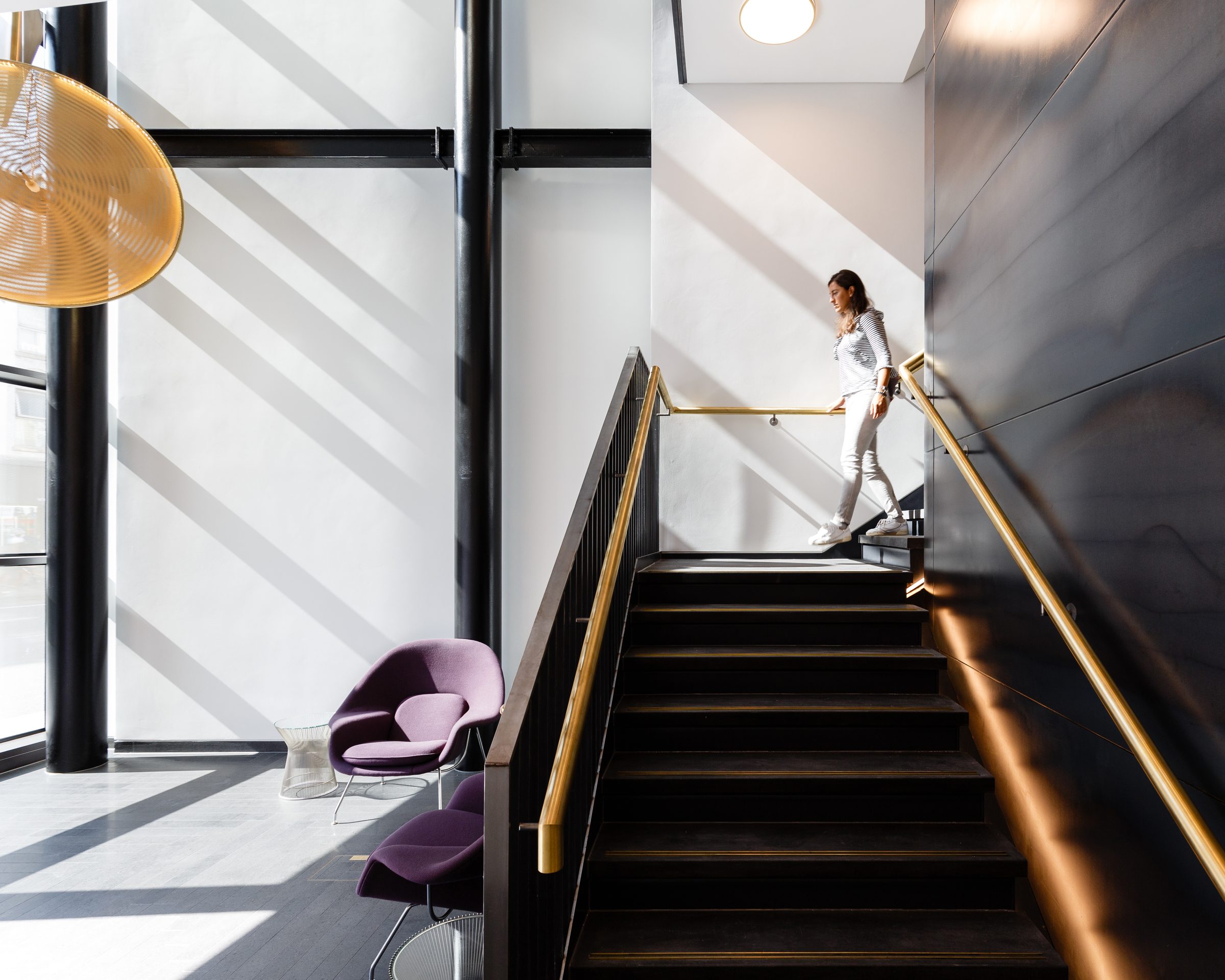 Stair with a brass handrail against a dark metal-clad wall, lit by daylight from the full-height glazing