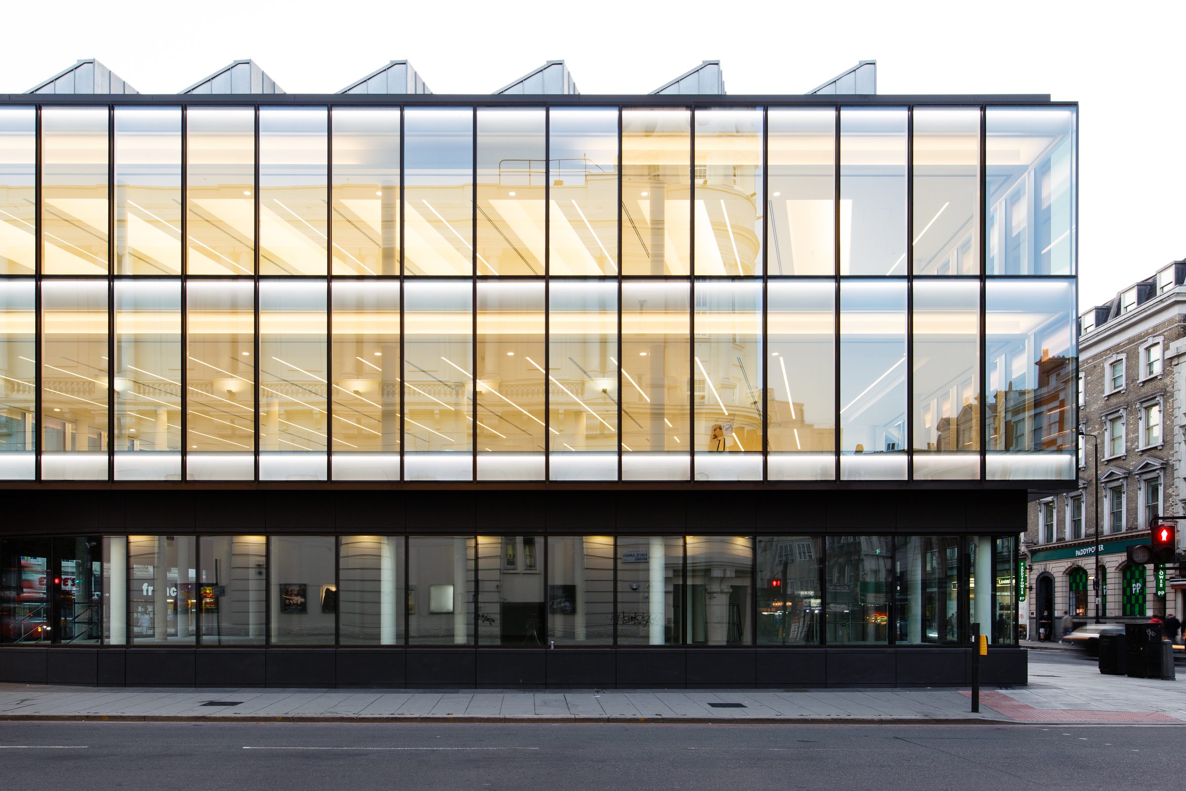 The glazed elevation lit from within at dusk, with the sawtooth roof profile above