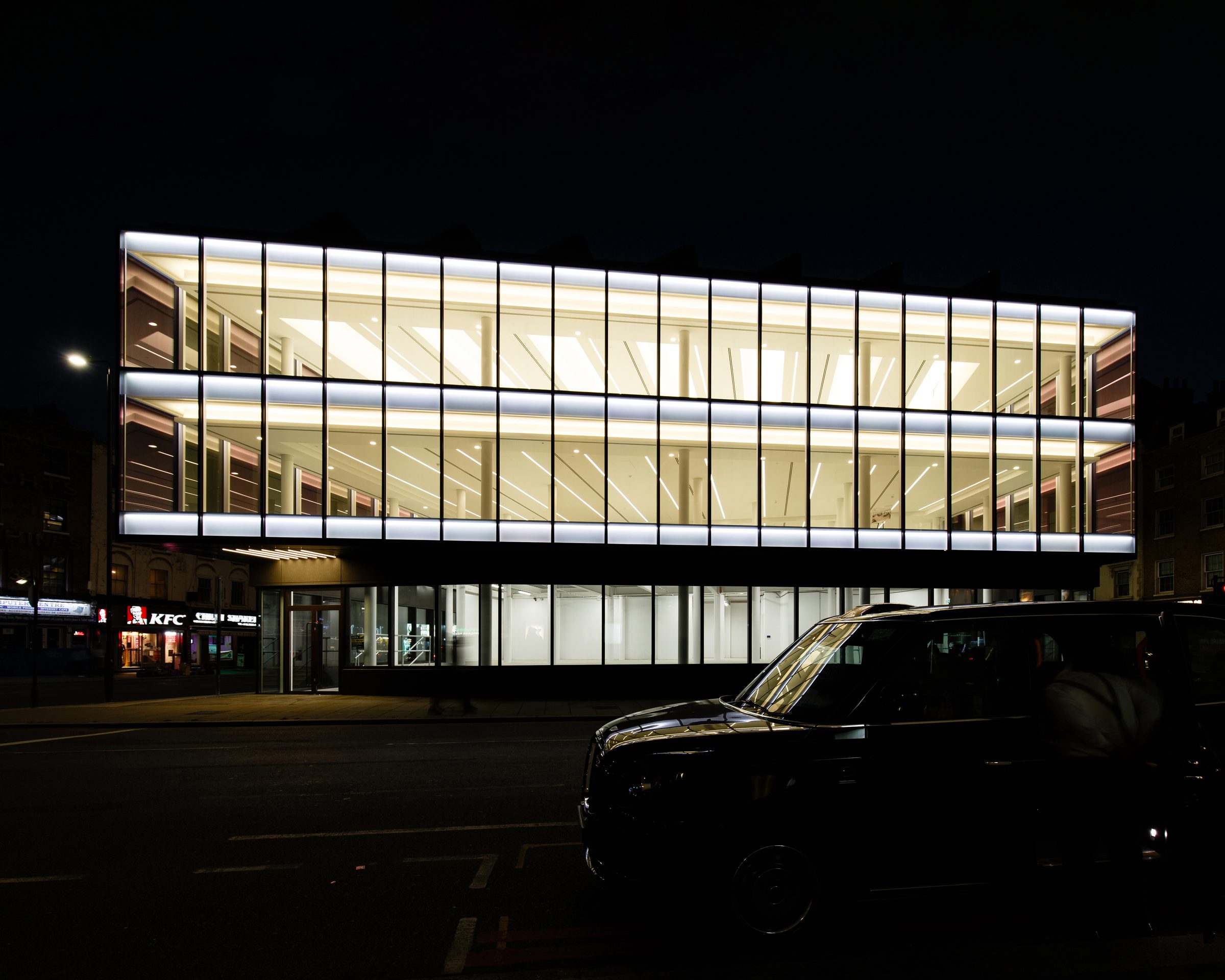 The illuminated glazed floors at night, with a black cab passing in the foreground