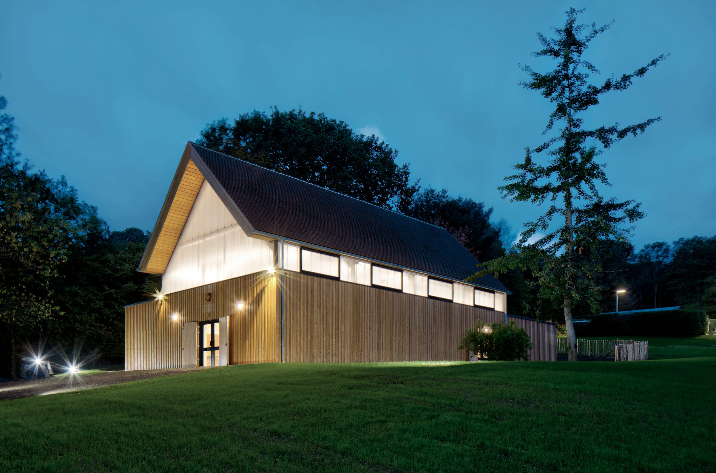 The timber-clad pavilion lit at dusk, with lawn and mature trees around it