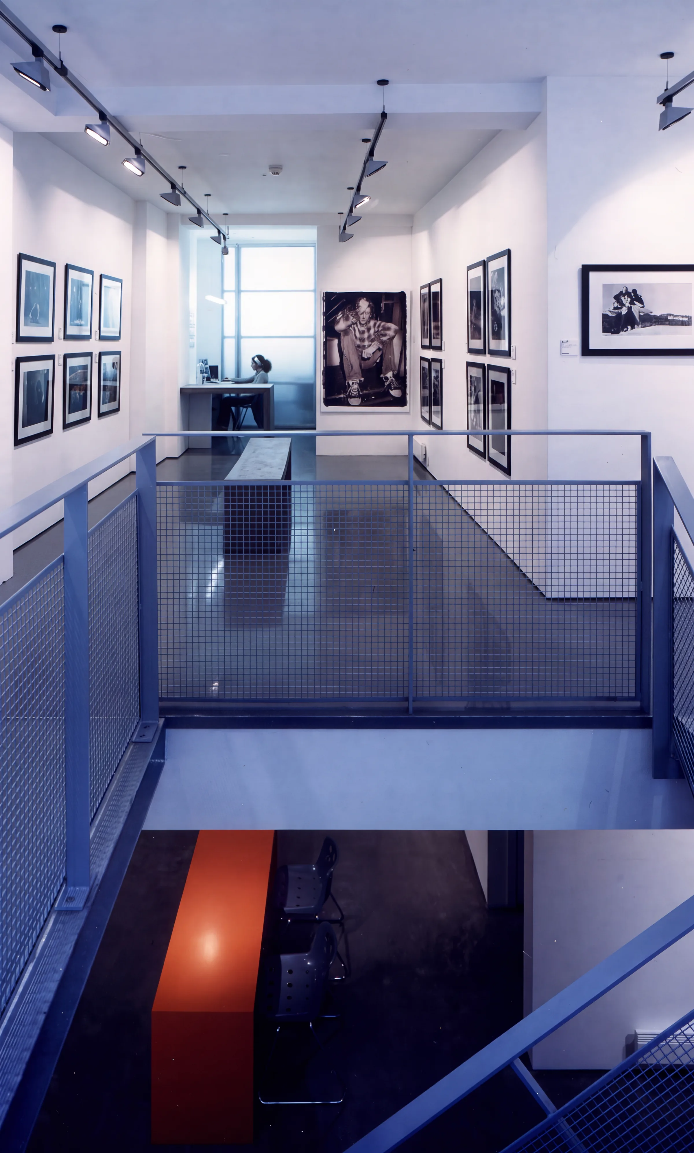 Looking down over the mezzanine, with track-lit photographs and a blue steel and mesh balustrade